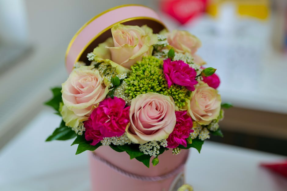 A round floral arrangement consisting of soft pink roses with full, layered petals, bright magenta carnations, and clusters of small green buds, all arranged in a pink cylindrical box with a silky pin
