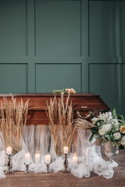 A collection of floral arrangements displayed in front of a polished wooden piano against a teal wall. The display includes tall, feathery beige pampas grass and white flowers arranged in a white cera