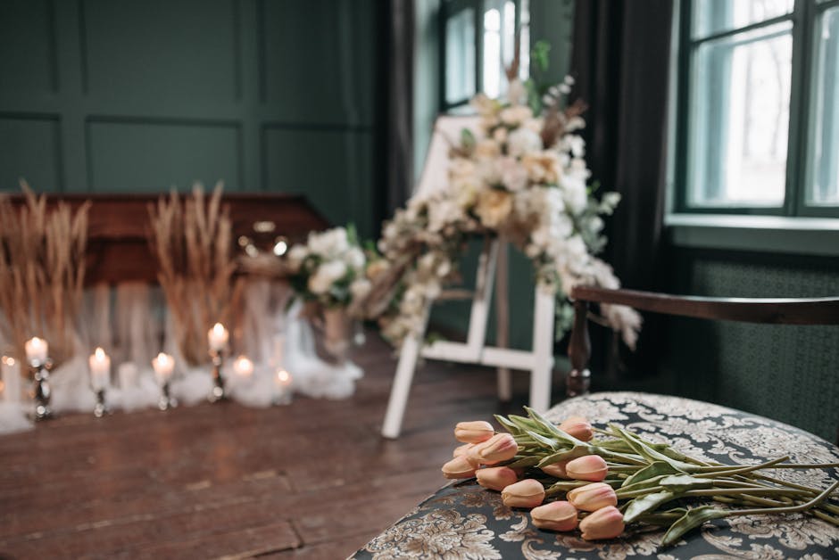 A floral arrangement of pale pink tulips with green stems and leaves is casually placed on an upholstered chair with a patterned fabric in a softly lit room. In the background, a large funeral wreath